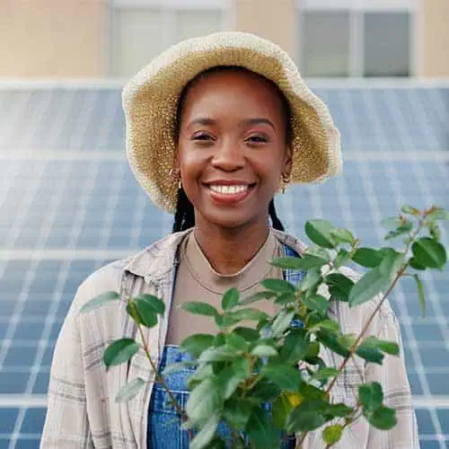Smiling woman outdoors with solar panels and green plants, promoting sustainable construction.